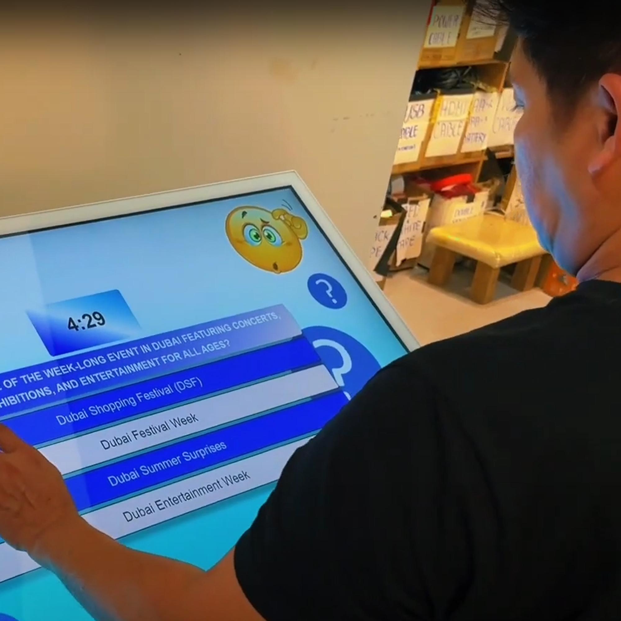 Touchscreen showing quiz results and a Dubai tower trivia question, with answer options and a person in a dark shirt standing beside the screen